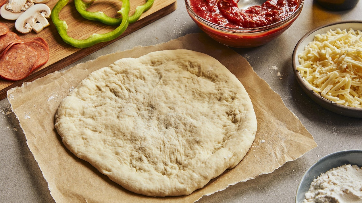 Une pâte à pizza déjà étirée est posée sur un papier parchemin. Autour, il y a une planche avec des légumes coupés et du pepperoni.