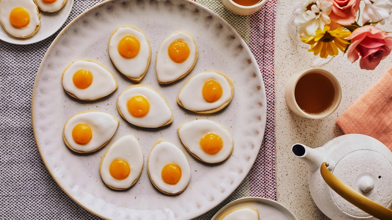 Dans une grande assiette de service, il y a plusieurs biscuits en forme d’œufs, avec une théière, des tasses de thé et des fleurs.