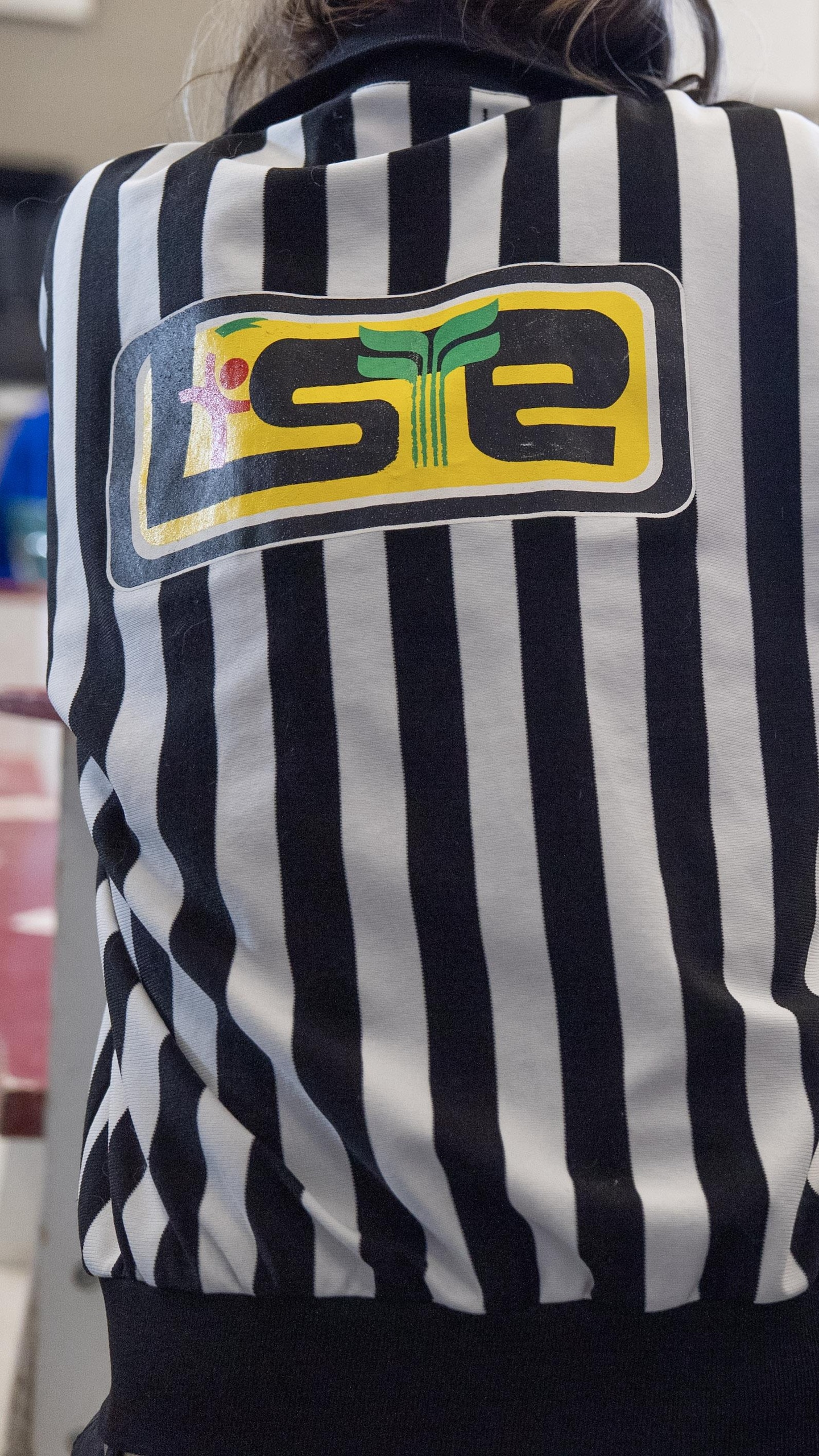 Vue de dos d'un arbitre portant le chandail classique à rayures verticales noires et blanches, avec le logo "LISTE" dans le dos.