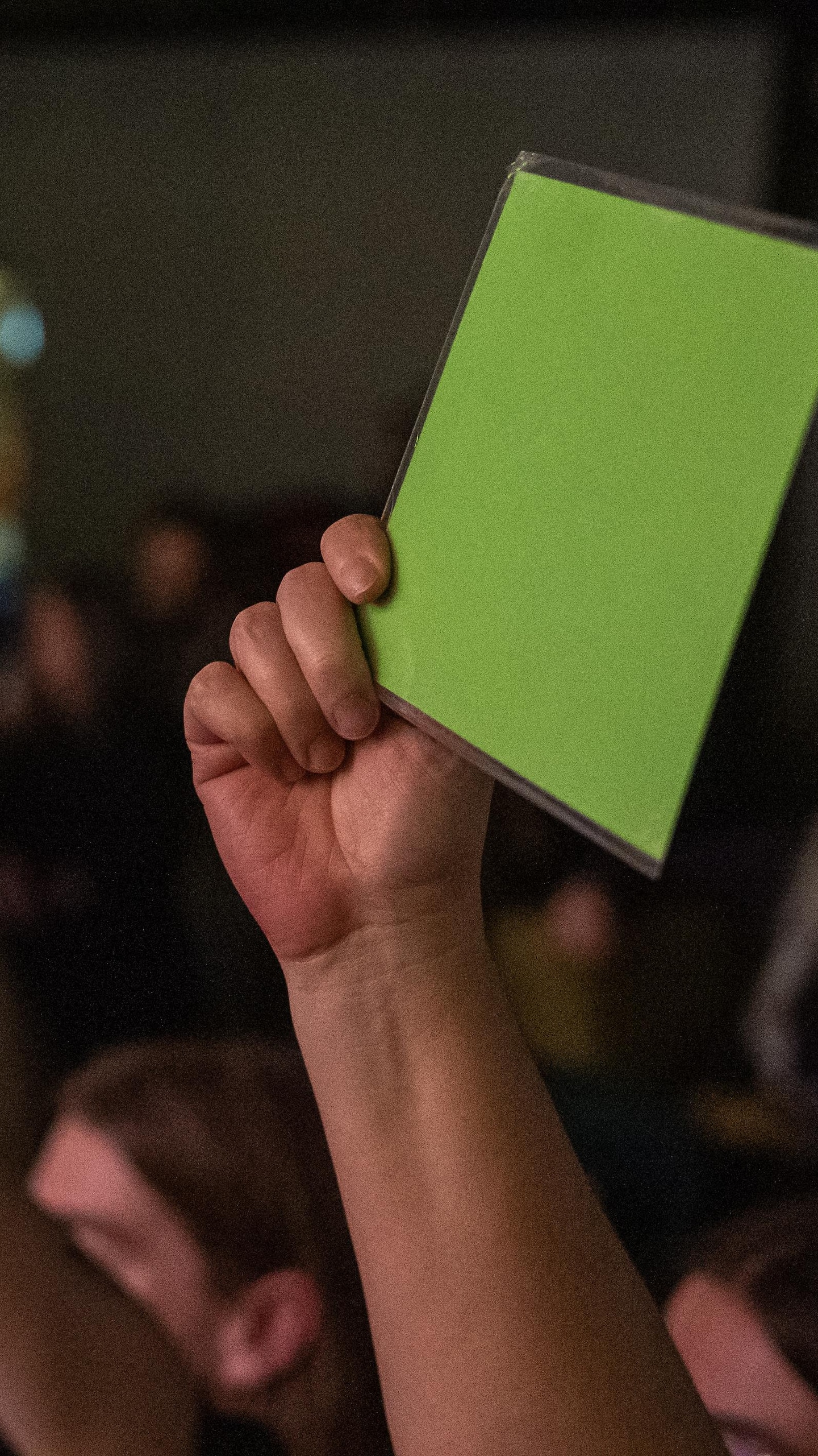 Gros plan sur une main brandissant un carton de vote vert vif devant une foule floue dans une salle de spectacle.
