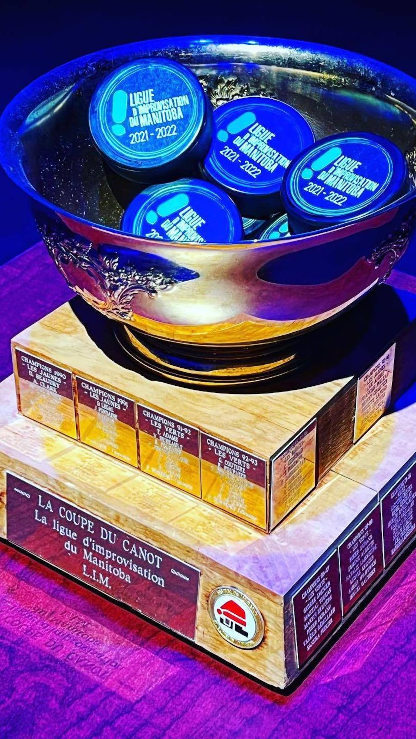 Une coupe argentée en forme de bol remplie de rondelles de hockey bleues marquées "LIM". Le trophée repose sur un socle en bois à plusieurs étages où sont fixées des plaques commémoratives.