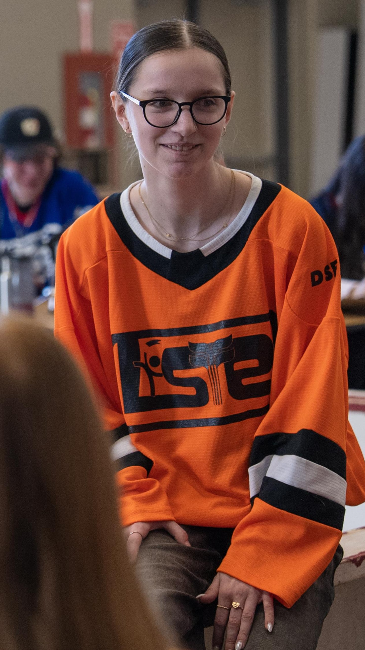 La jeune Gabrielle St. Pierre, portant des lunettes et un chandail orange, est assise sur le rebord en bois de la "patinoire" d'improvisation, regardant ses coéquipiers avec attention.