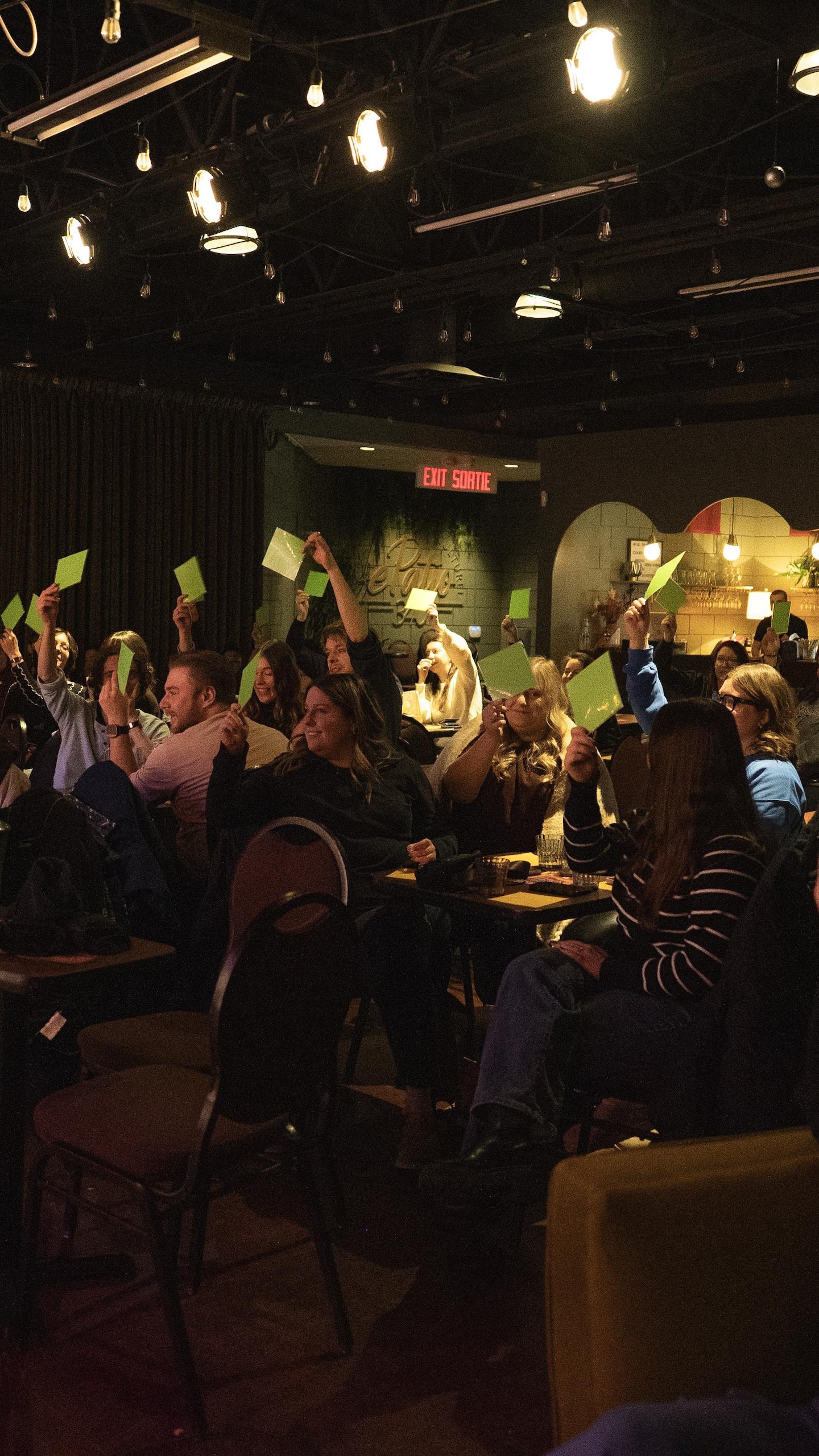 Une salle de spectacle bondée et sombre où de nombreux membres du public lèvent des cartons de vote colorés (verts et jaunes) pour choisir leur équipe préférée.