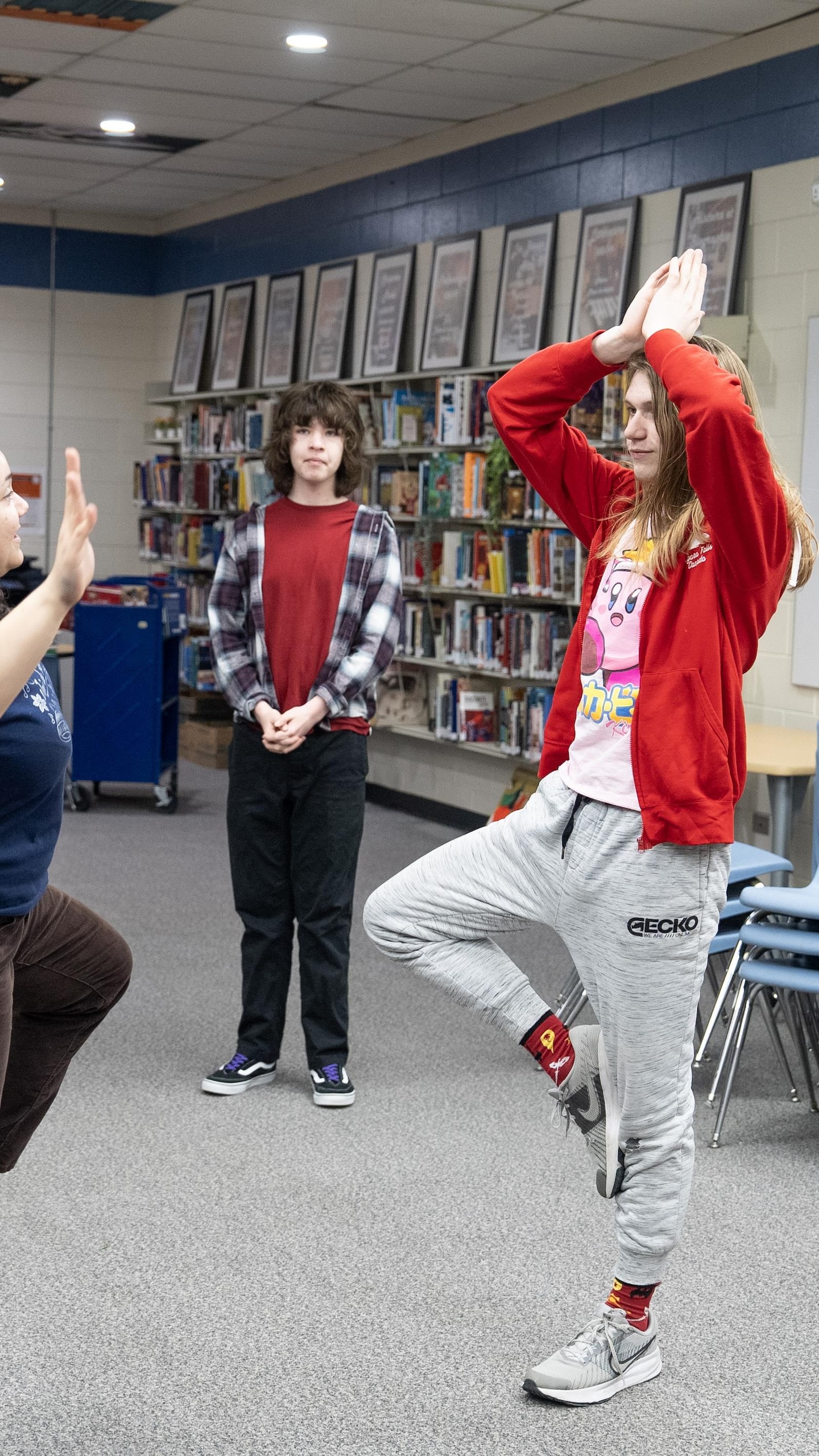 Zara Ramlal et un élève sont debout dans une bibliothèque, les bras levés dans des poses symétriques comme s'ils effectuaient un exercice de mime ou de miroir.