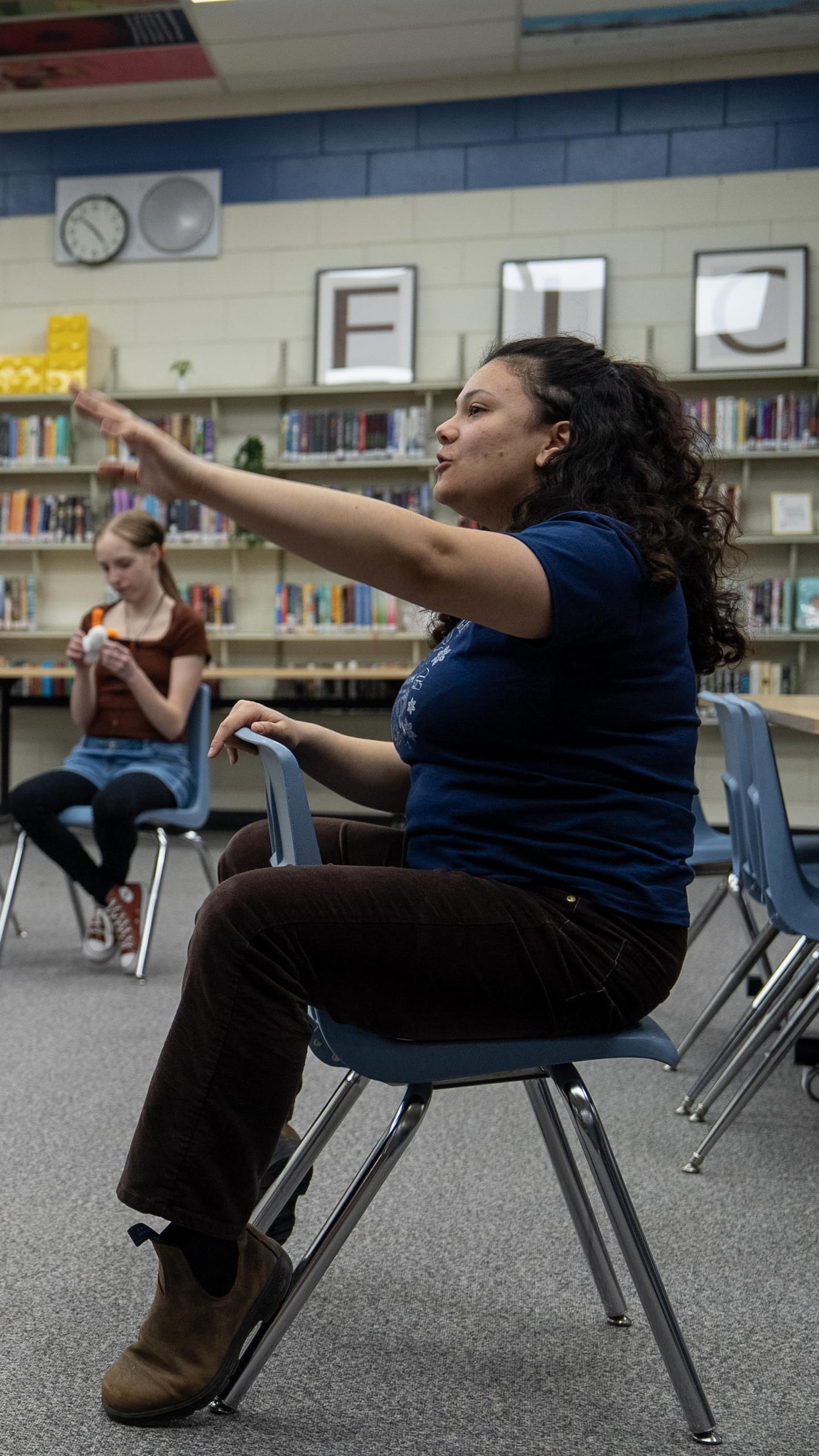 Zara Ramlal est assise dans une bibliothèque scolaire, faisant un geste de la main pour diriger un groupe de cinq adolescents assis en demi-cercle devant elle.