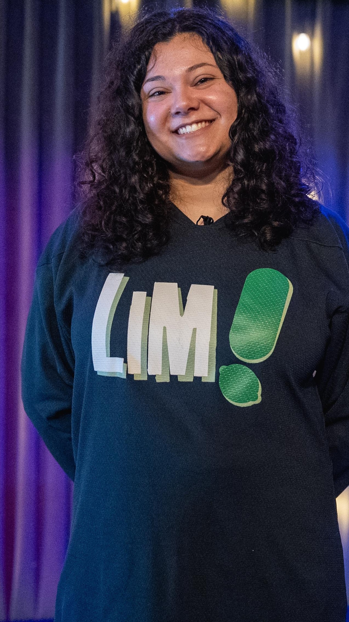 Portrait de l'improvisatrice Zara Ramlal, souriante, debout sur une scène devant un rideau violet illuminé de petites guirlandes lumineuses. Elle porte un chandail noir avec le logo "LIM".