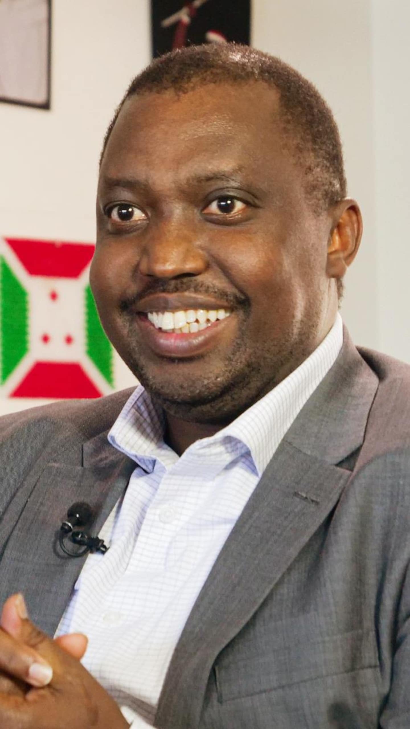 Portrait en plan moyen d'un homme noir souriant, vêtu d'un veston gris sur une chemise claire. Il a les mains jointes et se tient dans une pièce où l'on aperçoit des cadres de photos et un petit drapeau du Burundi au mur en arrière-plan.