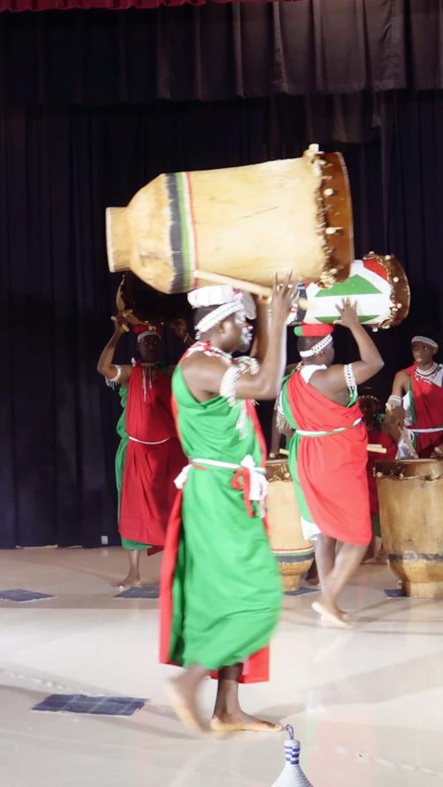 Vue d'ensemble sur une scène où plusieurs hommes en toges traditionnelles manipulent des tambours; trois d'entre eux marchent en portant leur instrument sur la tête, tandis que les autres sont alignés en arrière-plan.