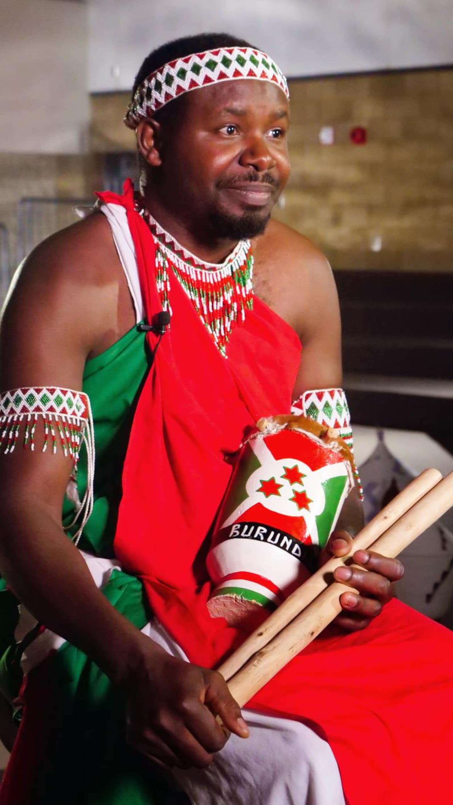 Un homme en costume traditionnel rouge et vert est assis de trois-quarts face, tenant deux baguettes de bois épaisses entre ses mains. Un mini-tambour décoré est posé sur ses genoux.