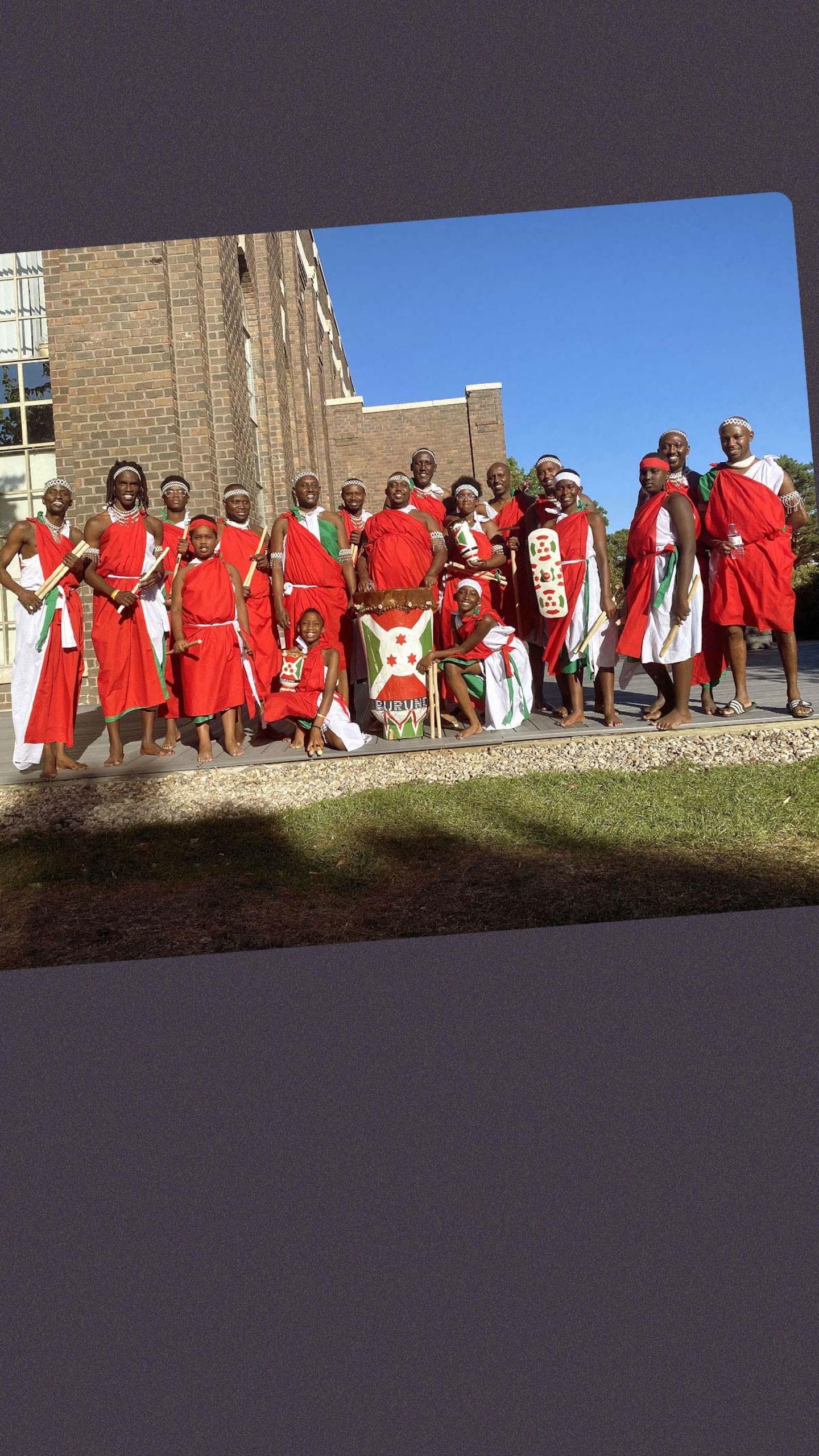Une vingtaine de personnes de tous âges, vêtues de tenues traditionnelles rouges et blanches, posent côte à côte à l'extérieur devant un bâtiment en briques sous un ciel bleu. Un grand tambour décoré est posé au centre du groupe.