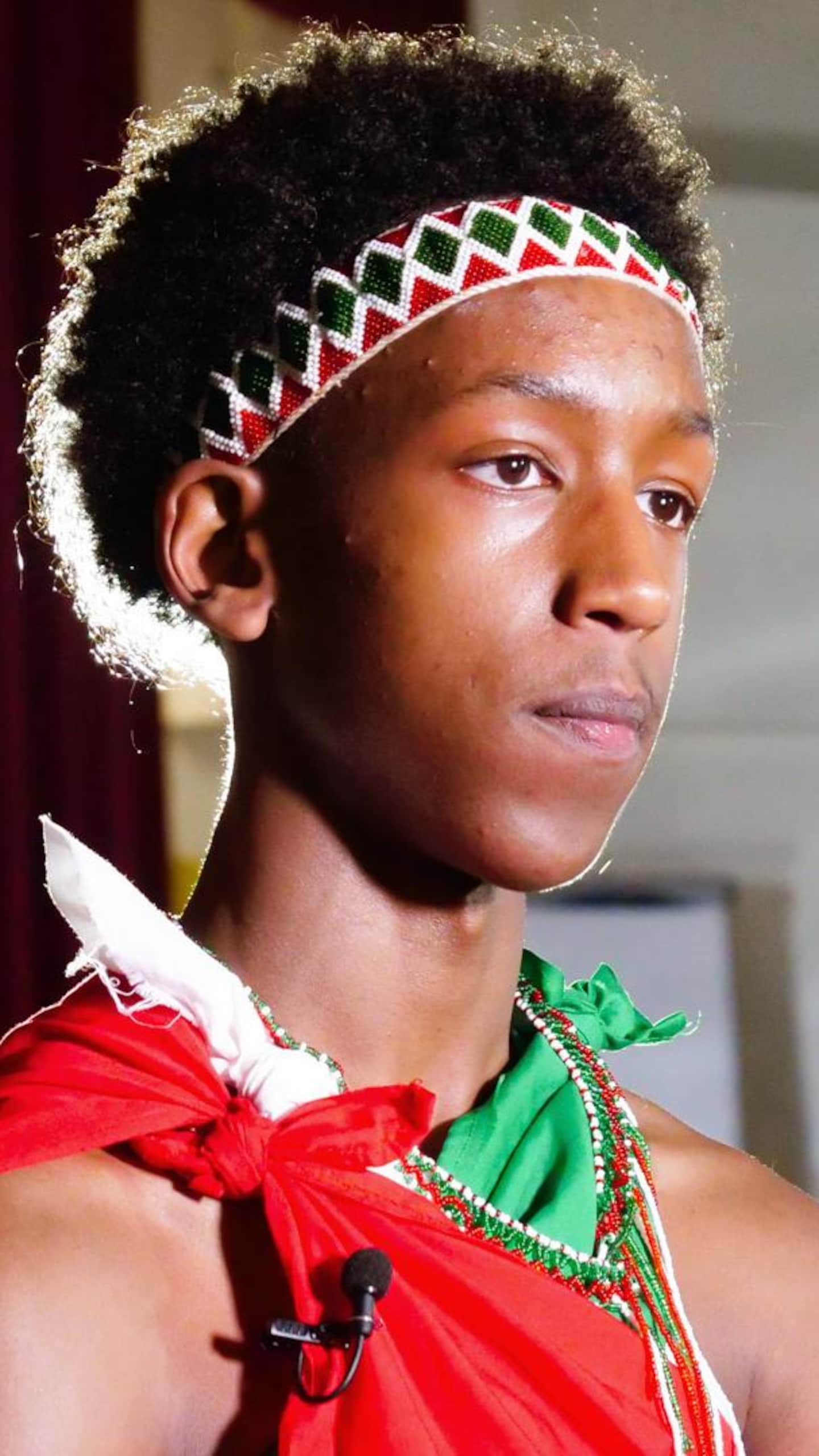 Portrait de profil d'un jeune homme portant un bandeau perlé blanc, rouge et vert, vêtu d'une toge rouge, se tenant sur une scène de théâtre.
