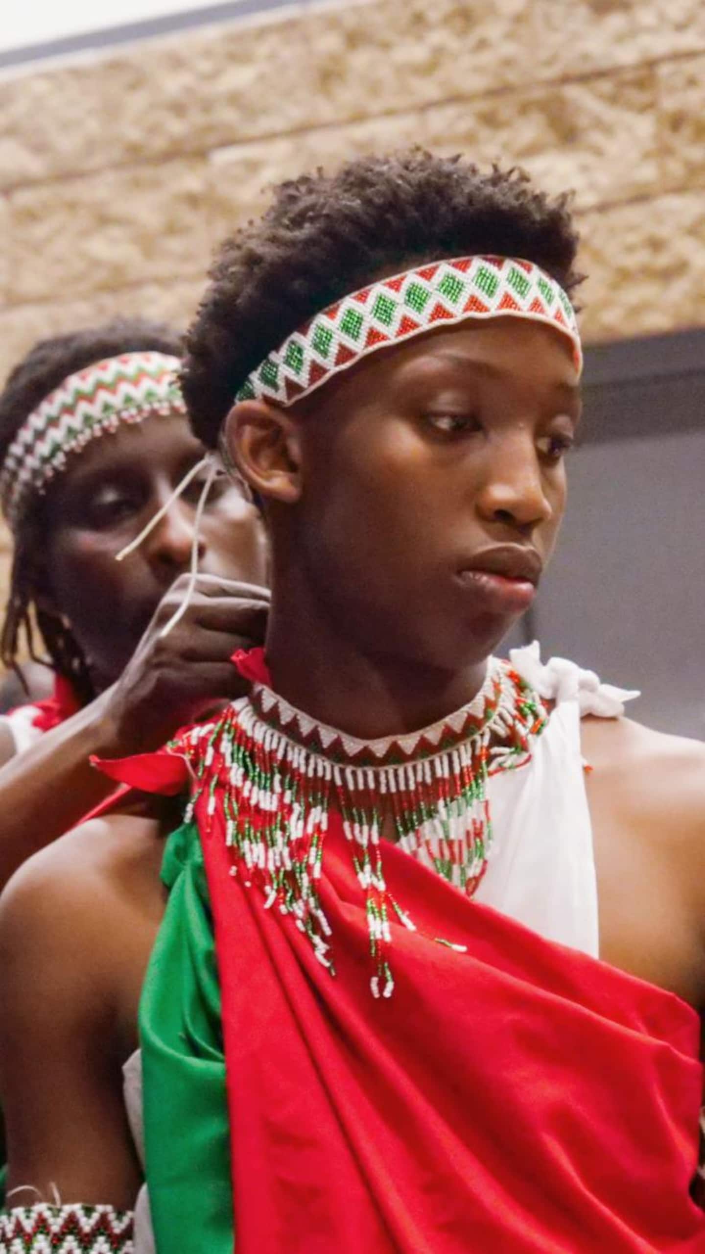 Gros plan sur un jeune homme de profil portant une toge rouge et verte. Un collier de perles blanches imposant entoure son cou, et une personne en arrière-plan ajuste le bandeau perlé noué autour de sa tête.