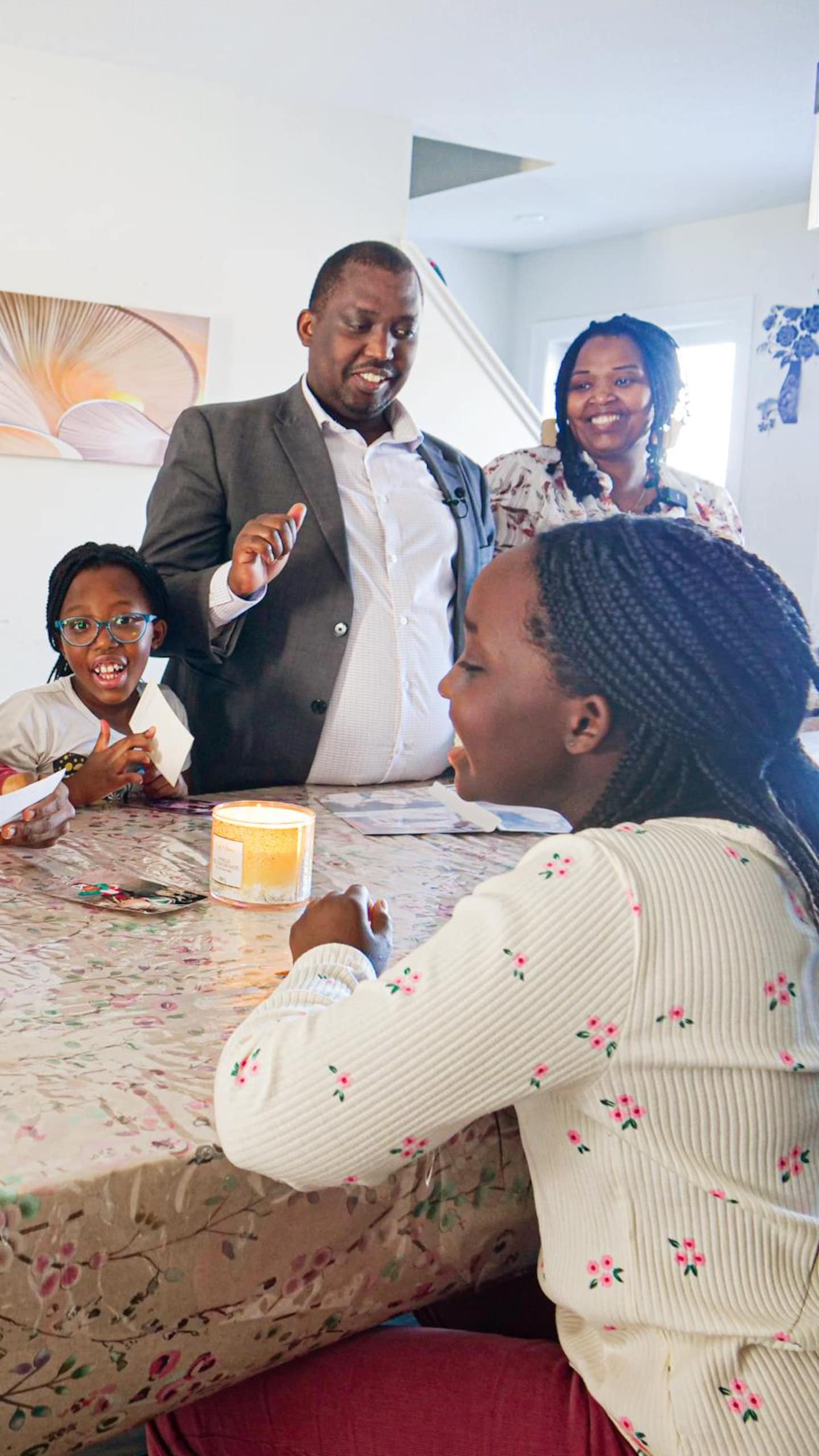 Une famille de six personnes est réunie à l'intérieur autour d'une grande table en bois. Un homme en veston est debout au centre de la pièce, tandis qu'une femme et quatre enfants sont assis autour de la table.