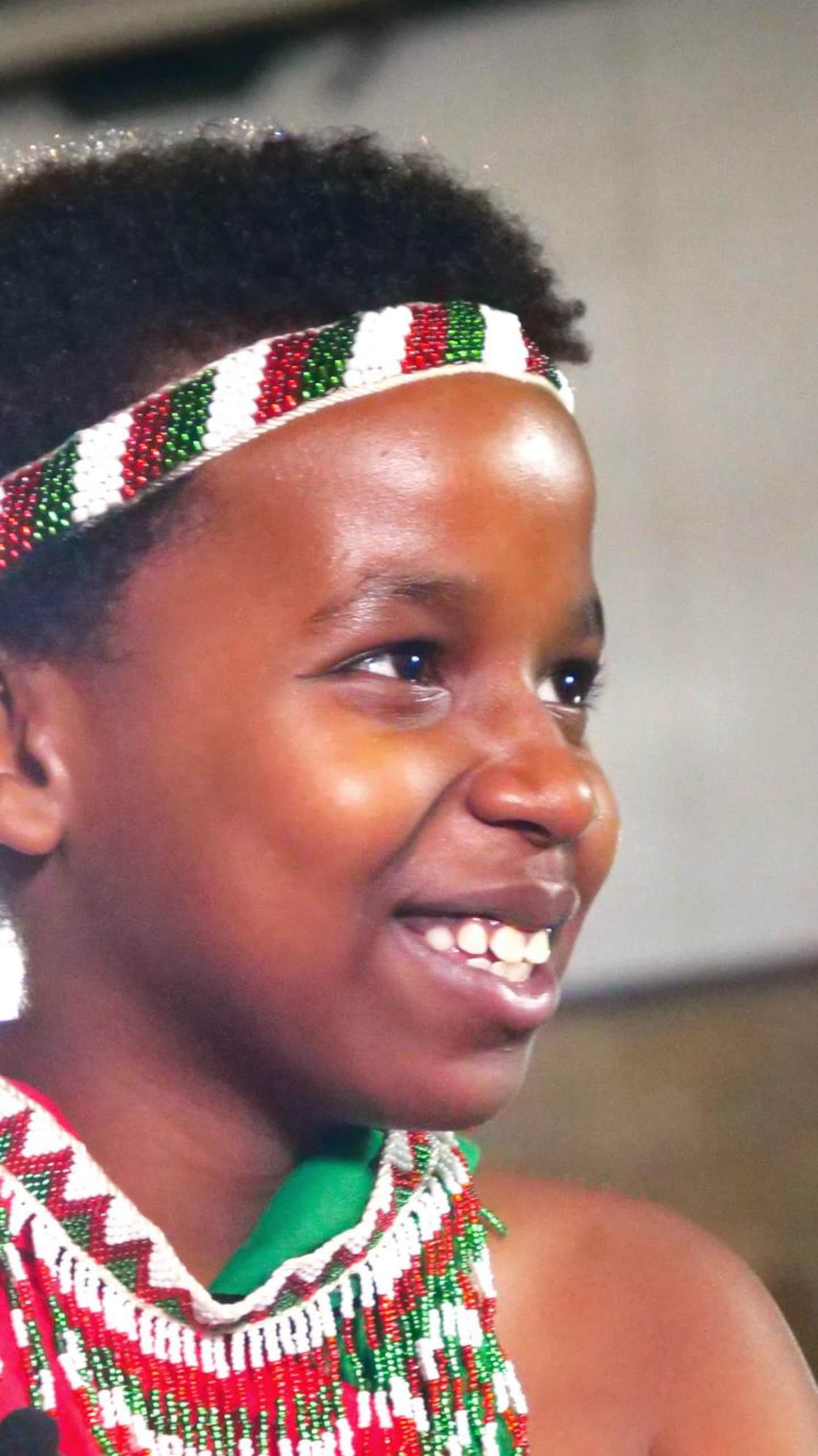 Portrait rapproché d'un jeune garçon noir souriant de profil, portant un bandeau perlé et un collier traditionnel blanc, rouge et vert.