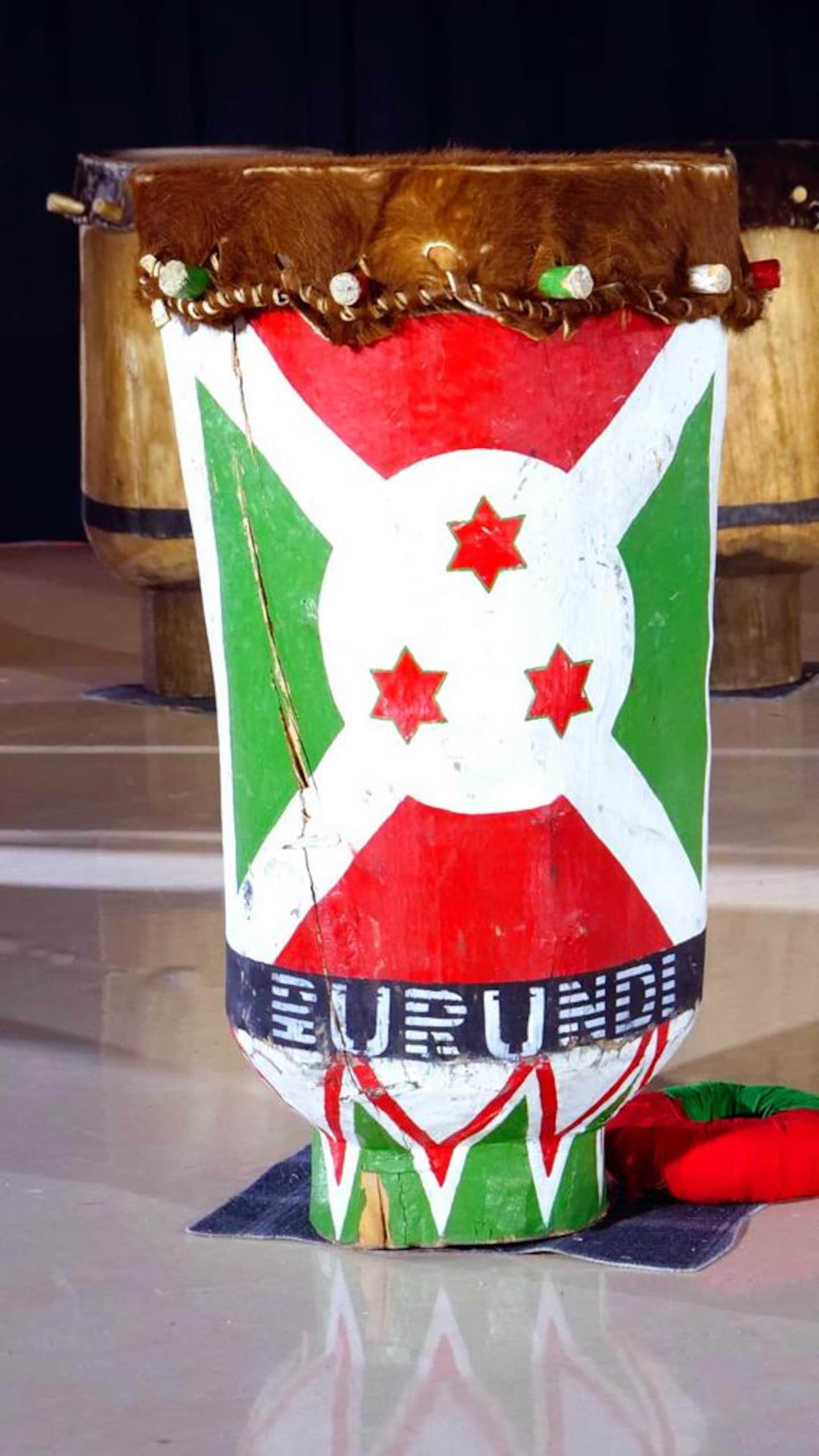 Un grand tambour en bois, décoré d'un panneau central peint en rouge, blanc et vert avec trois étoiles au milieu, est isolé au centre d'une scène vide, devant d'autres tambours alignés à l'arrière-plan.