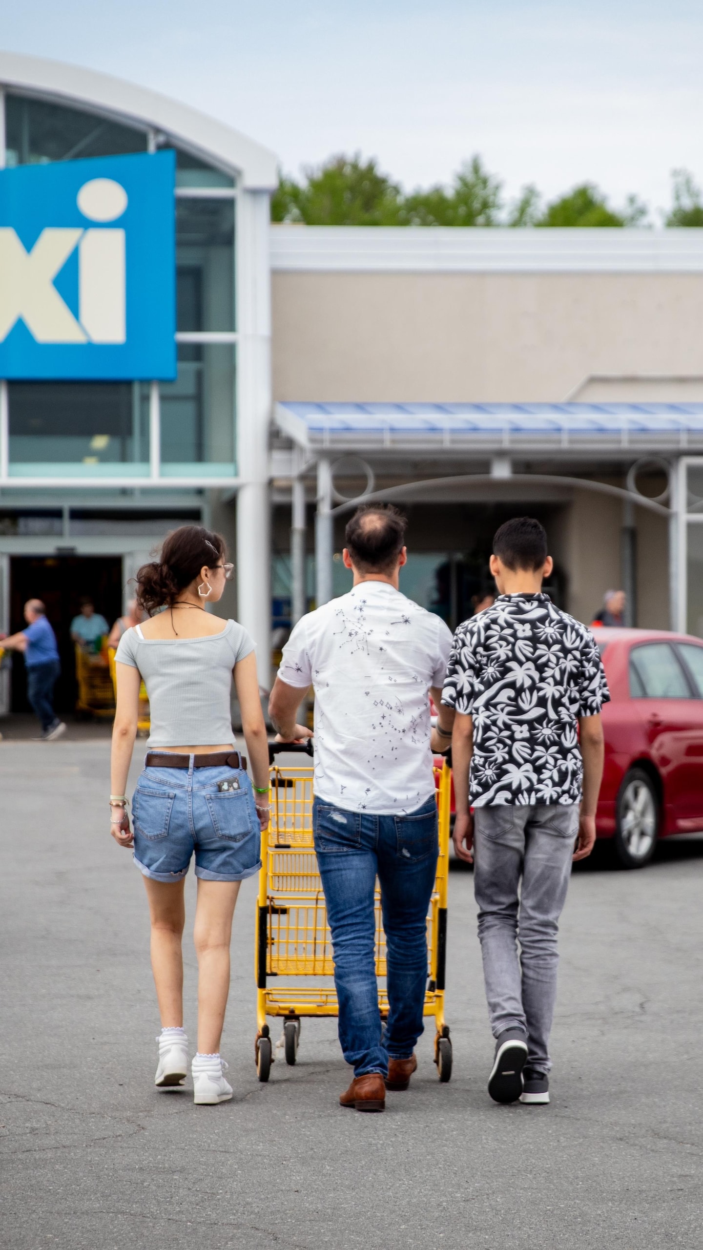 Trois personnes marchant, vues de dos, poussent un panier d'épicerie dans un stationnement en direction d'un supermarché Maxi.