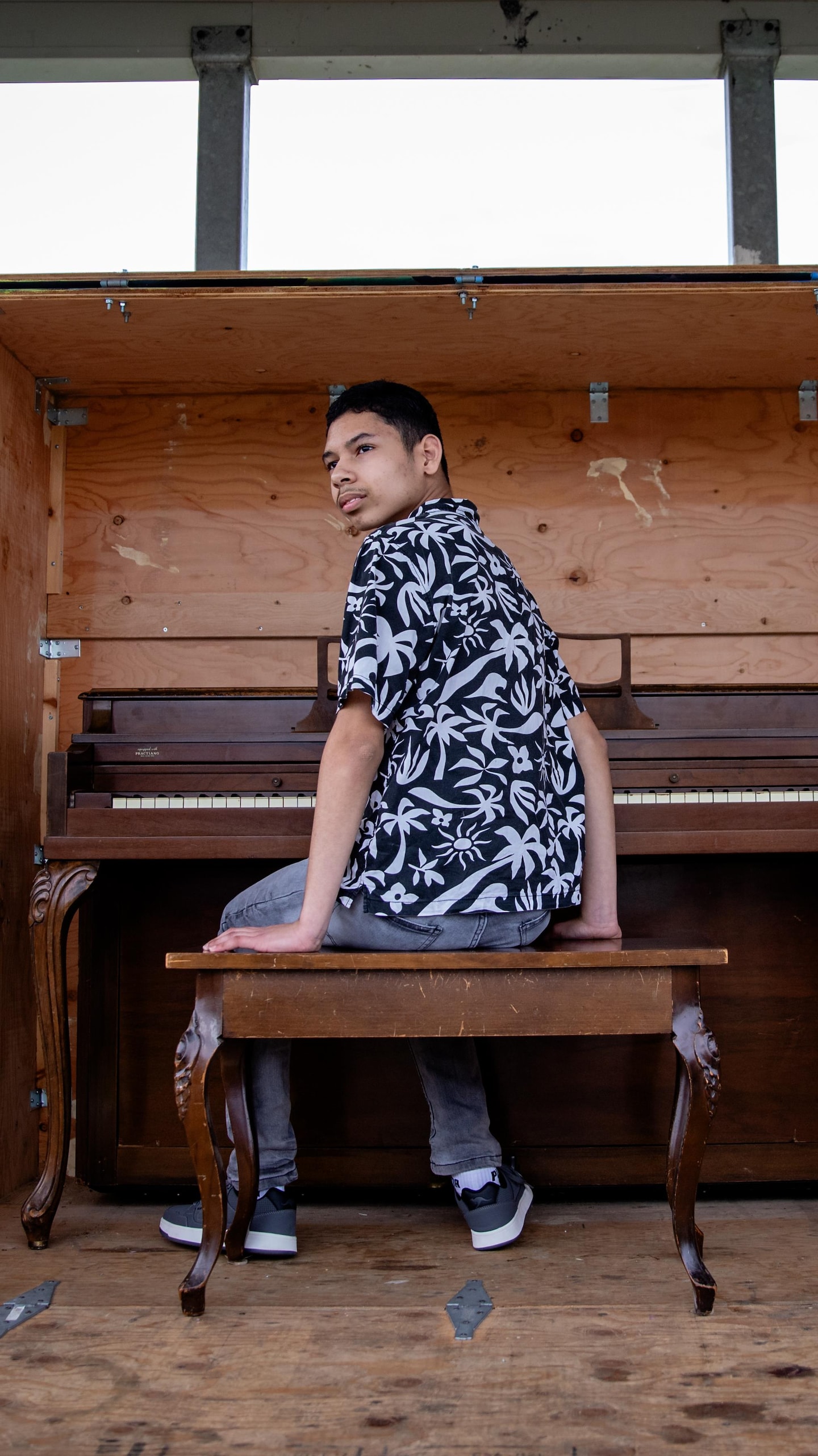 Un garçon est assis sur le banc d'un piano public contenu dans une boîte en bois, sous un gazébo. 