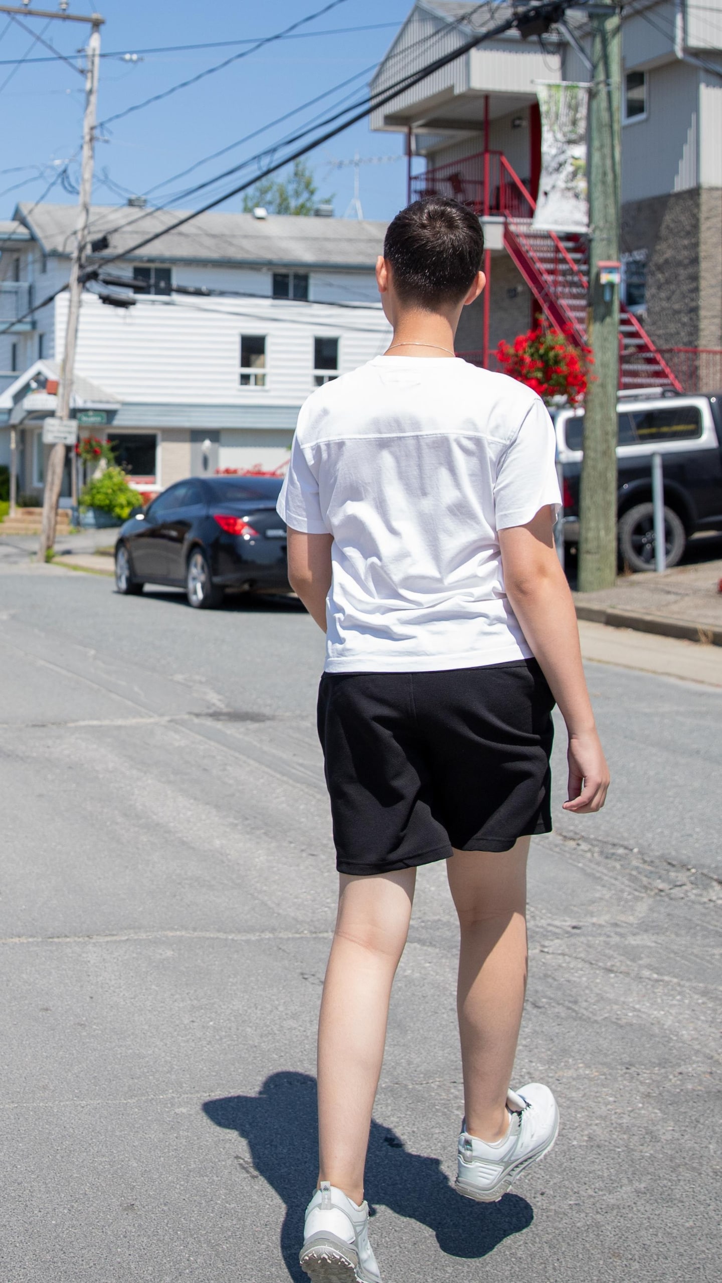 Un jeune homme en t-shirt blanc et en short noir marche, vu de dos, au milieu d'une rue résidentielle bordée de maisons.