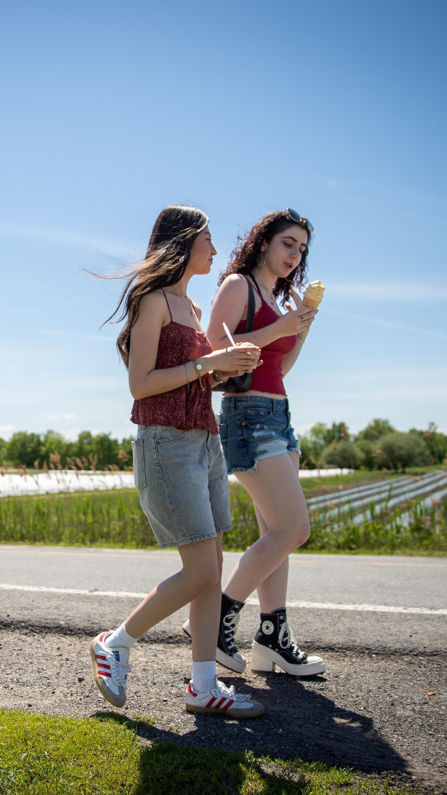 Deux jeunes femmes marchent côte à côte le long d'une route rurale en mangeant une crème glacée par une journée ensoleillée.