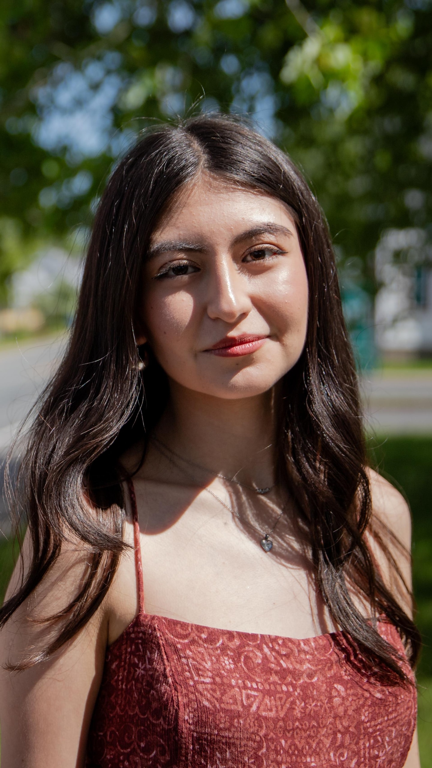 Une jeune femme aux cheveux bruns pose en extérieur près d'une rue bordée d'arbres et de maisons.