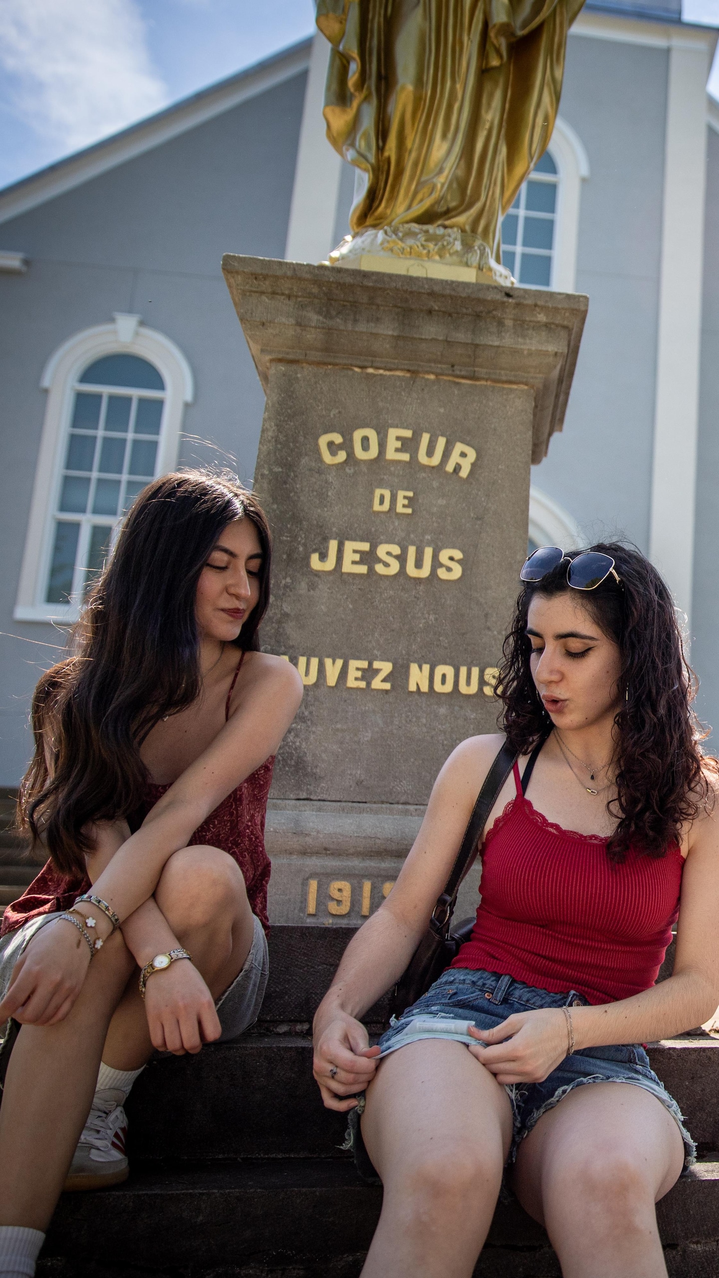 Deux jeunes femmes discutent, assises sur des marches, au pied d'un monument dédié au Cœur de Jésus devant une église.