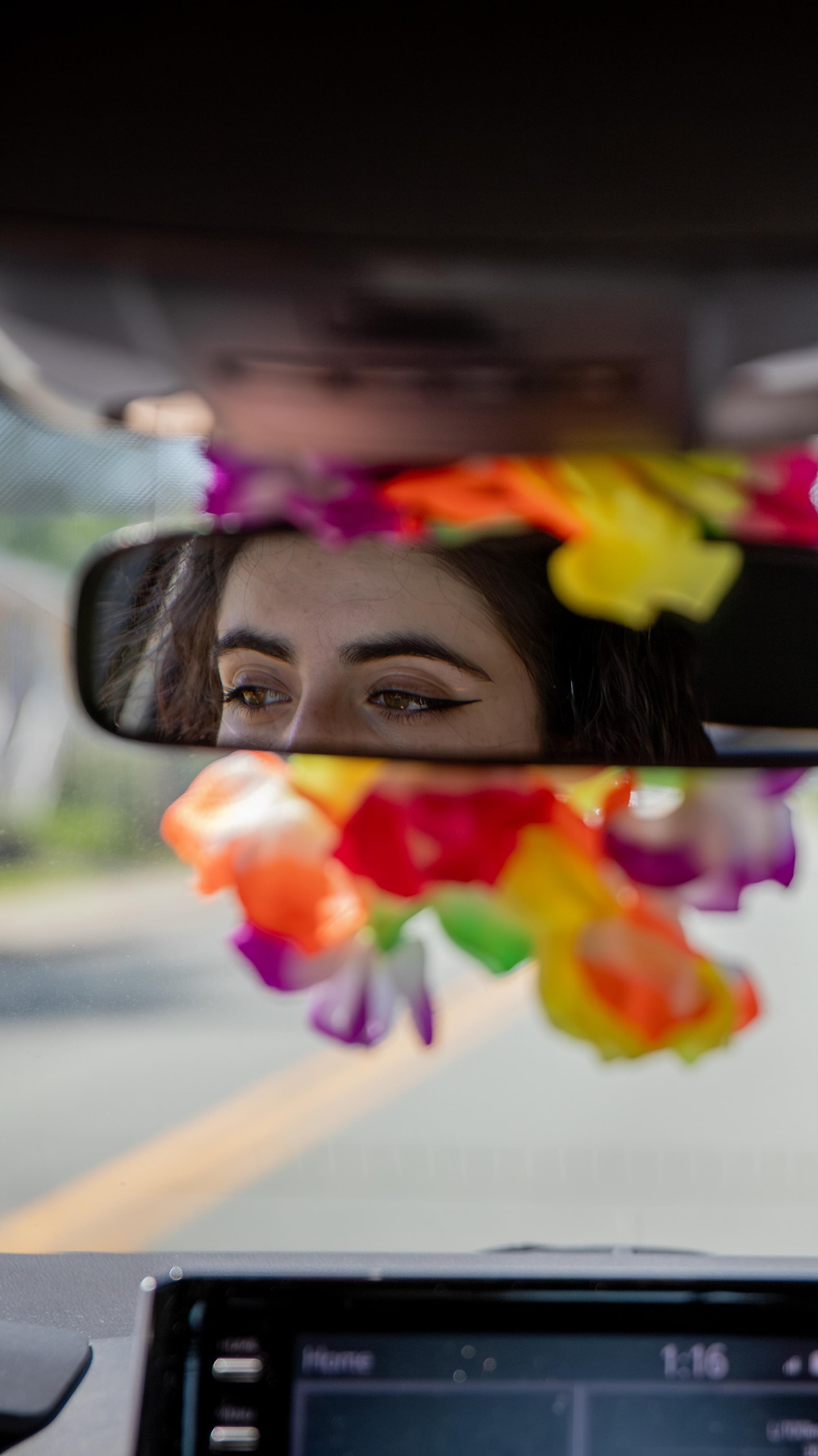 Le reflet des yeux d’une personne regardant la route apparaît dans le rétroviseur d’une voiture, entouré d'une guirlande de fleurs.