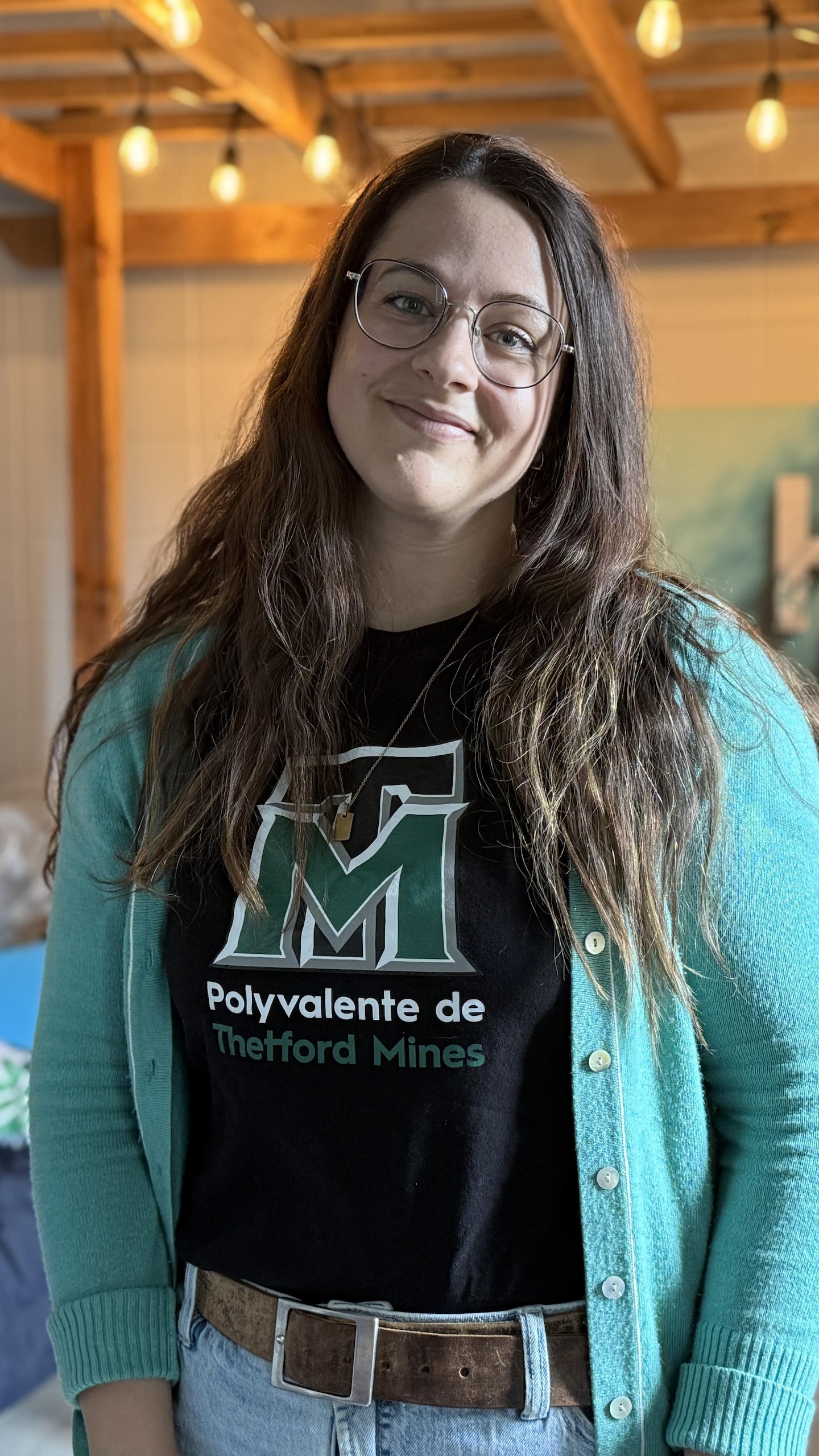 Une femme souriante à lunettes porte un chandail noir marqué « Polyvalente de Thetford Mines » sous un cardigan vert, dans une pièce éclairée.
