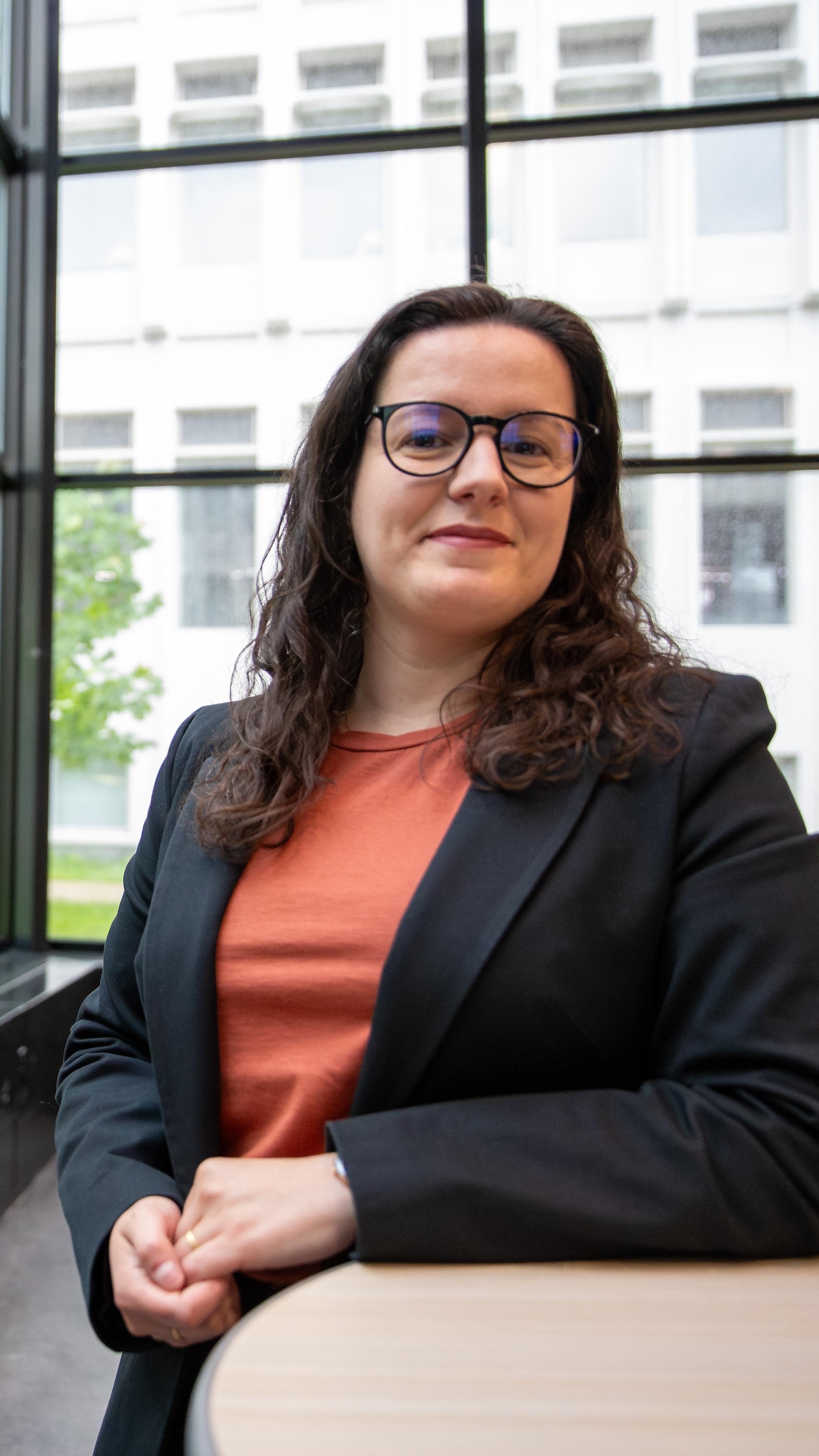 Une femme portant des lunettes et un veston noir s’appuie sur une table devant une grande baie vitrée.