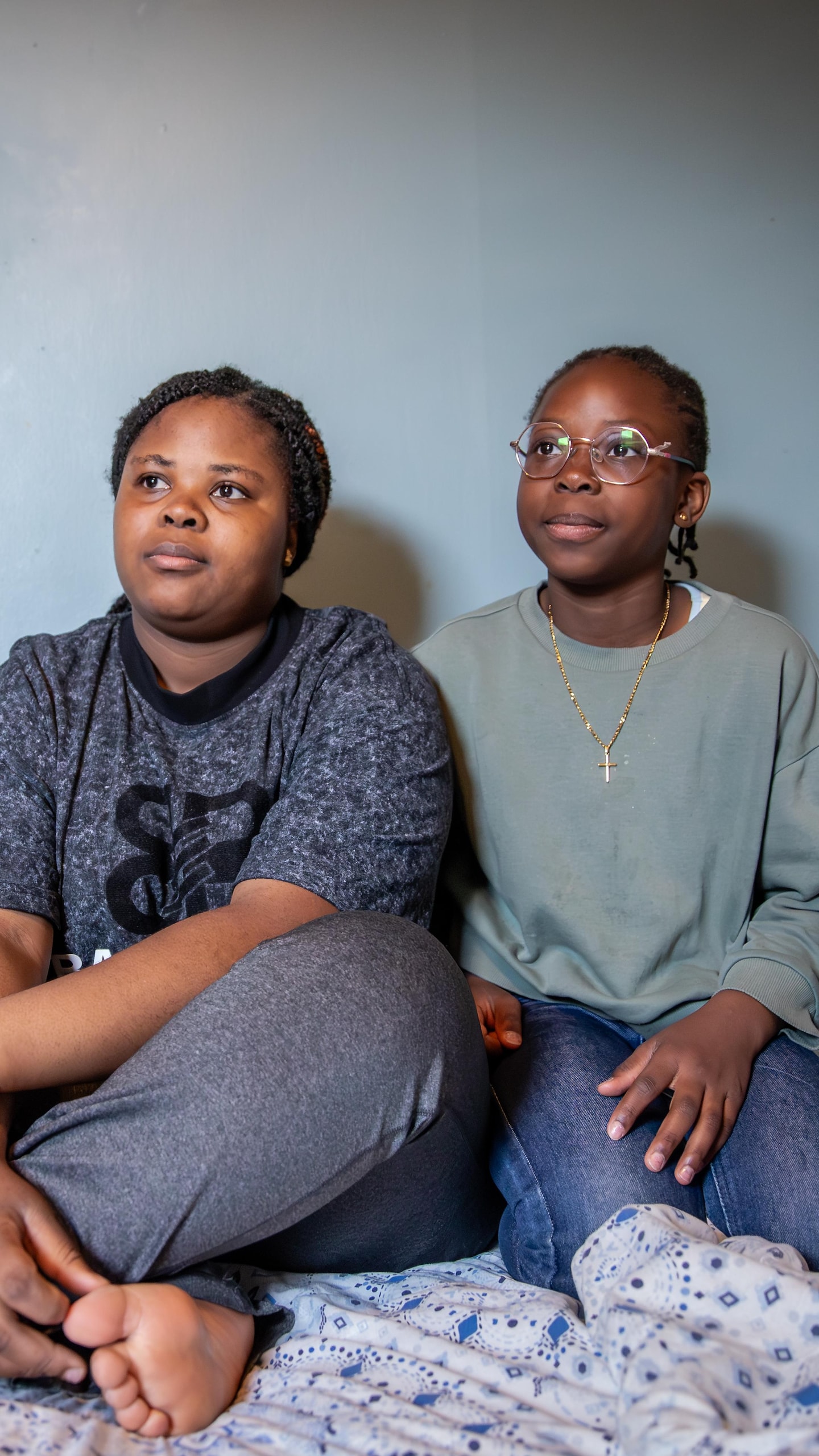 Une jeune femme et une fillette sont assises côte à côte sur un lit dans une chambre aux murs bleus.