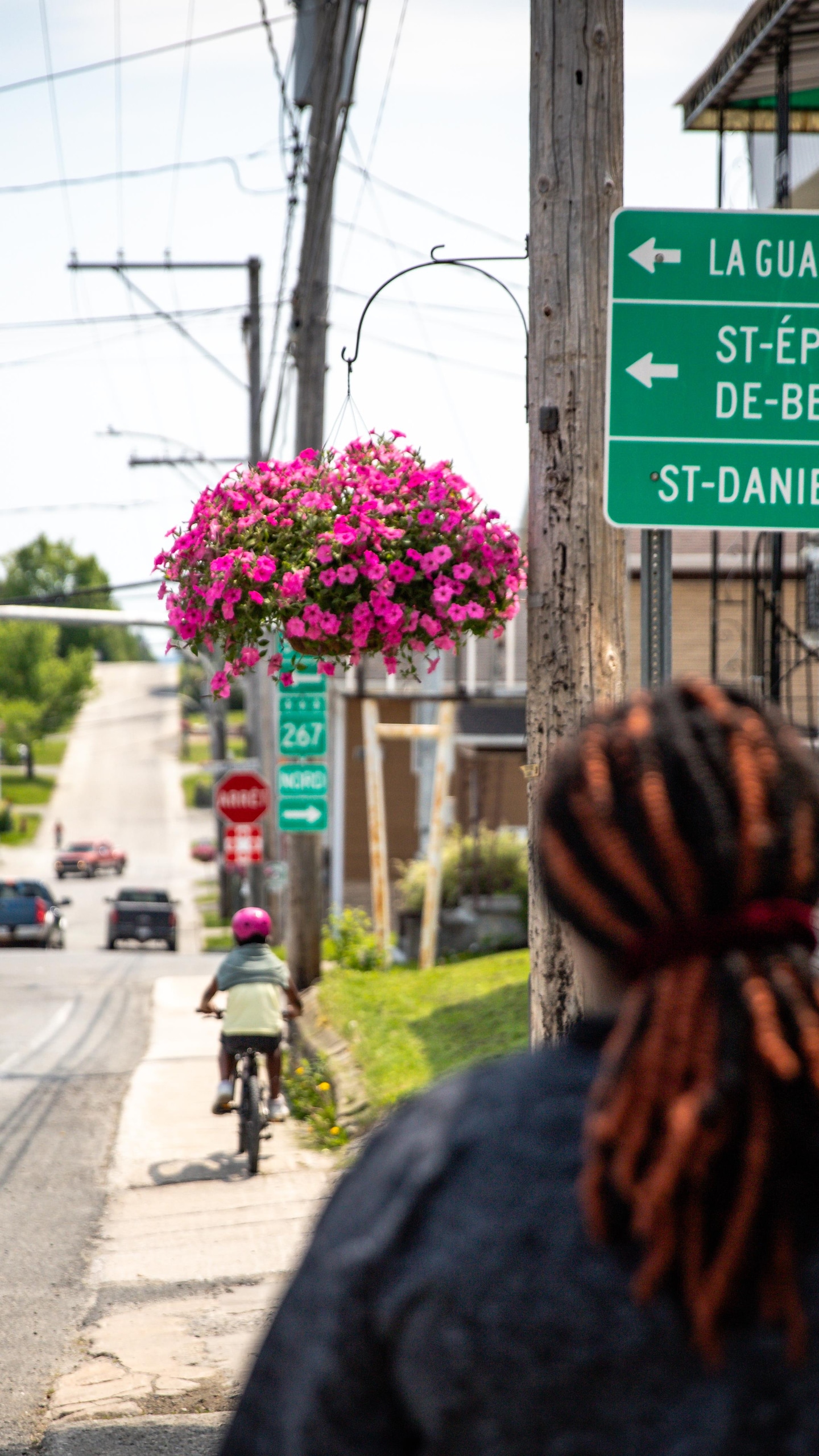 Une personne vue de dos regarde un panneau routier indiquant des directions, près d'un panier suspendu de fleurs roses. En avant d'elle, une petite fille fait du vélo.