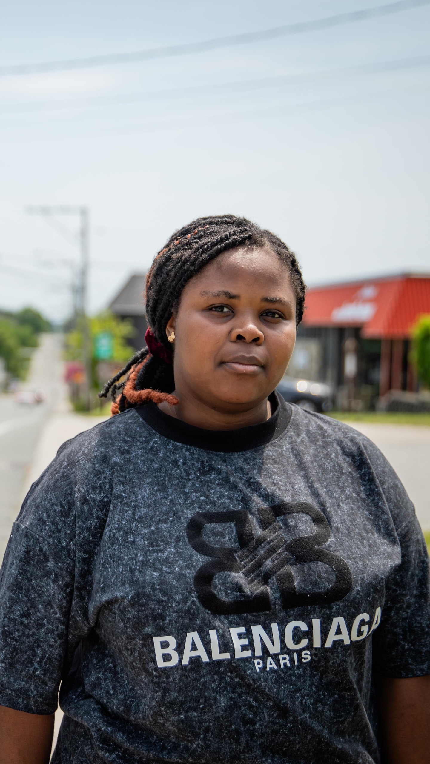 Une femme vêtue d'un t-shirt Balenciaga se tient en bordure d'une route déserte traversant un village.
