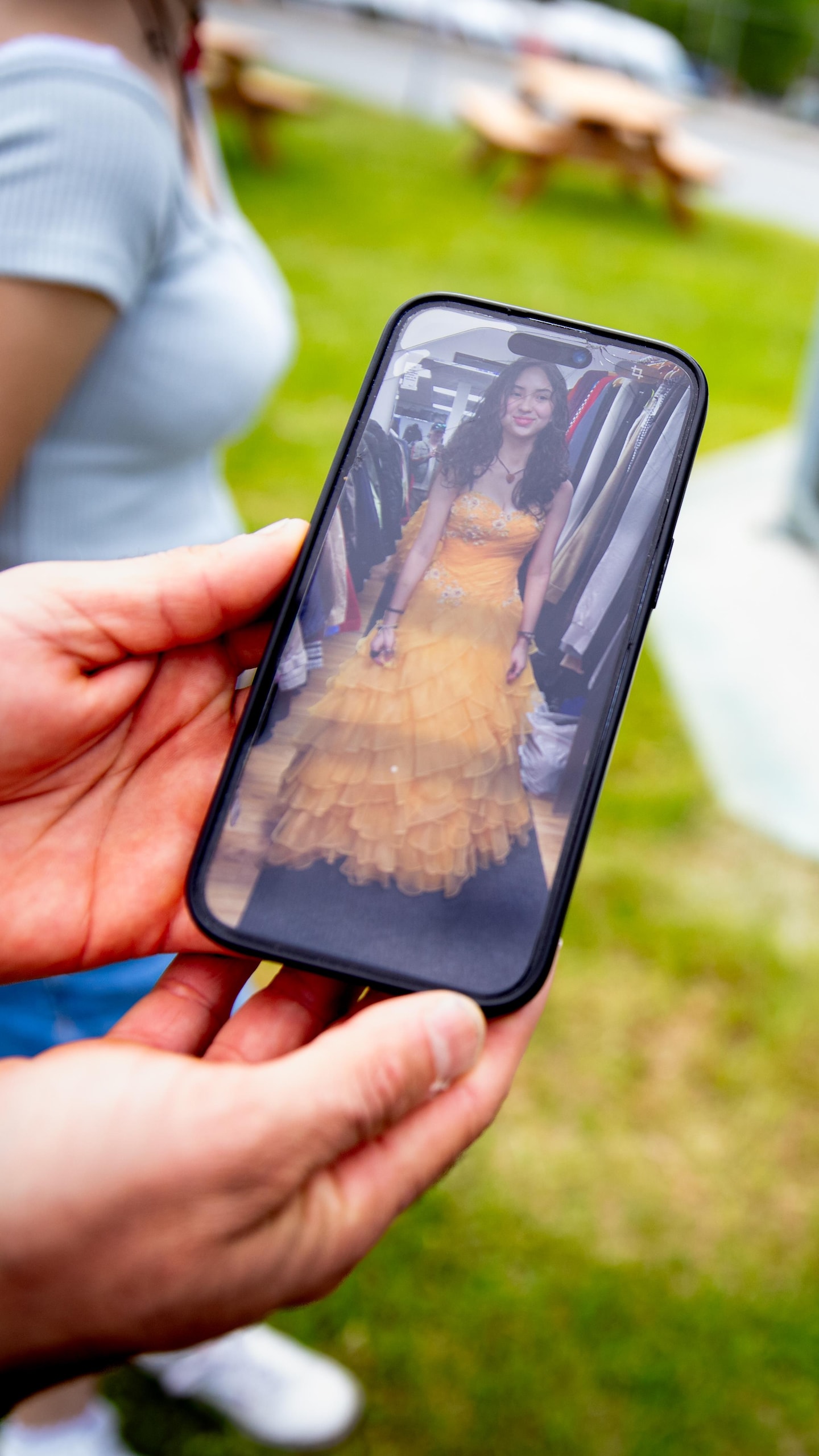 Des mains tiennent un téléphone affichant une jeune femme en robe de bal jaune, près d'un gazébo en extérieur.