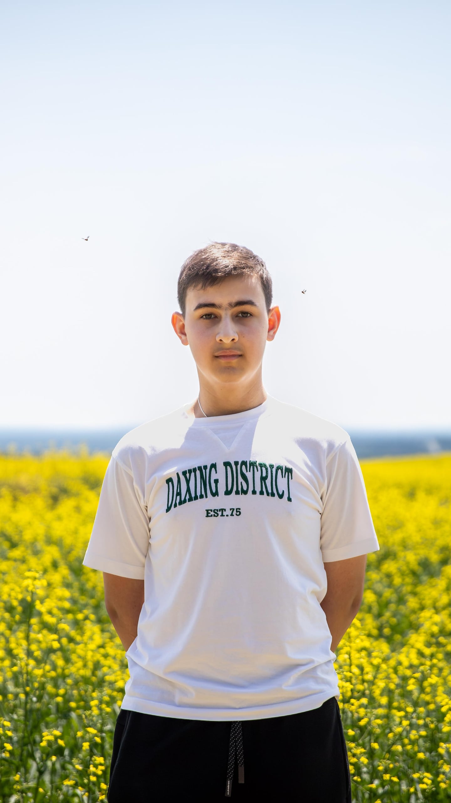 Un jeune homme regarde l'objectif, les mains dans le dos, devant un champ de fleurs jaunes. Deux mouches tournent autour de son visage. 