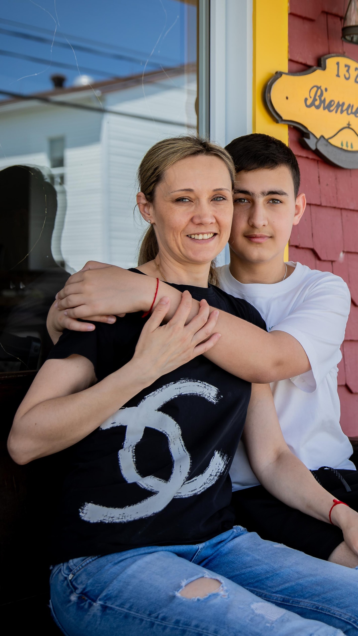 Une femme et un jeune homme sourient, assis sur un banc devant l'entrée d'un bâtiment au mur rouge portant l'enseigne « Bienvenue ».