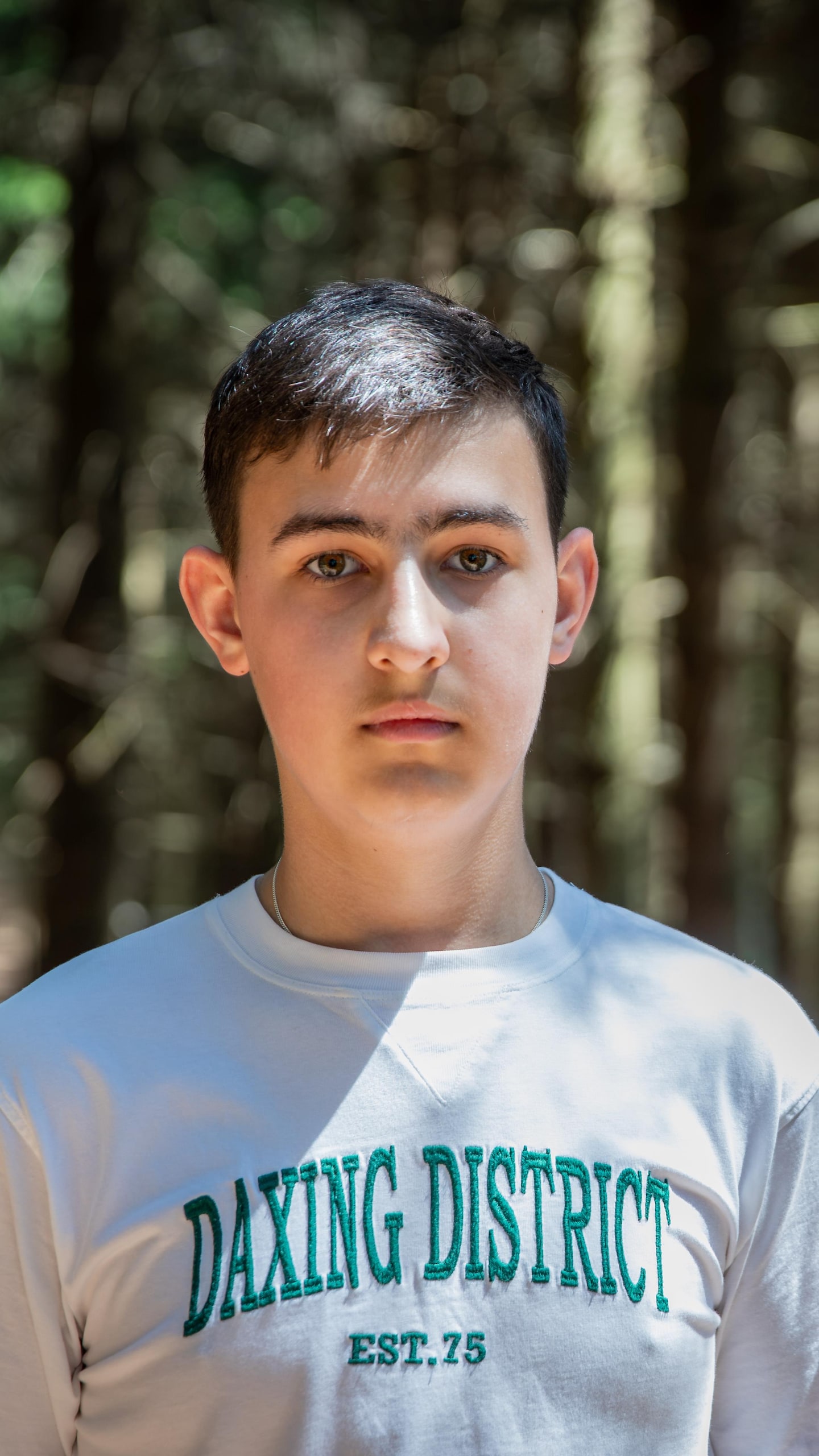 Un jeune homme portant un t-shirt blanc marqué « Daxing District » se tient debout dans une forêt.