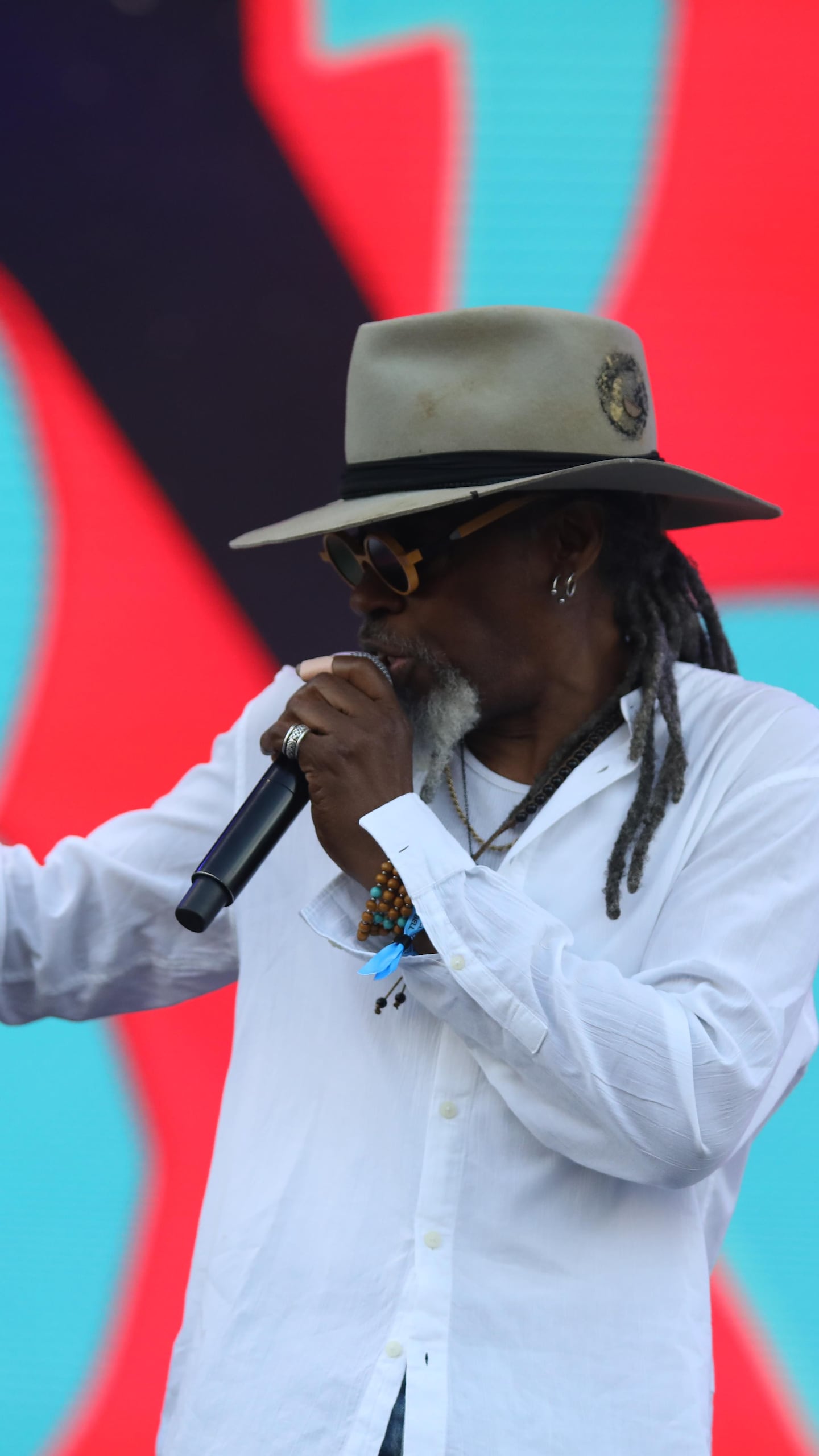 Lunettes de soleil au visage et chapeau sur la tête, Alan Prater chante en faisant un geste de la main.