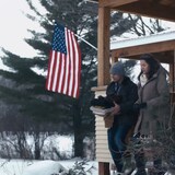 un homme et une femme sortent d'une maison, en hiver. Un drapeau américain est visible à l'arrière-plan.