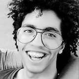 Portrait en noir et blanc d'Achraf Thimoumi, un jeune homme portant des lunettes de vision, souriant pour la caméra. Il a des broches et les cheveux bouclés.