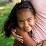 Une jeune fille afro-latino regarde la caméra et sourit. Elle est enlacée par une personne adulte, dont les bras sont visibles. 