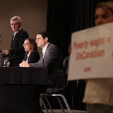 Air Canada officials speaking as a flight attendant holds a sign saying 'Poverty wages = UnCanadian'.