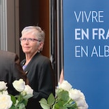 Une bannière sur laquelle se lit «Vivre en français en Alberta», à droite, avec la députée Annie McKitrick qui écoute un interlocuteur, vu de dos, avec des roses blanches au bas de l'image.