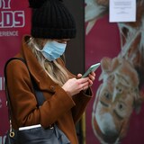 Une femme, coiffée d'une tuque en laine et le visage recouvert d'un masque médical, est penchée sur son téléphone cellulaire.