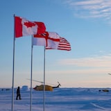 D'immenses drapeaux du Canada et des États-Unis, de même que le pavillon britannique White Enseign flottent au vent près de deux hélicoptères au sol dans l'Arctique.