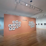 Esp: Vista de una sala de exposición de museo con una gran pared naranja que muestra fotografías enmarcadas y el texto "RECUERDO".
*
Fr: Vue d'une salle d'exposition avec un grand mur orange affichant des photos encadrées et le texte « RECUERDO ».