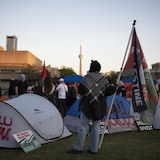 Une personne portant un keffieh et tenant un drapeau palestinien est debout devant des tentes. La Tour CN est à l'horizon.