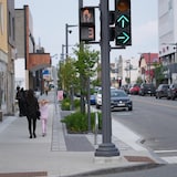 Des passants, dont une femme et une petite fille marchant le trottoir, ainsi que des véhicules stationnés de chaque côté de la rue.
