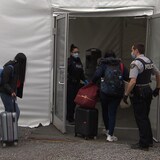 Trois jeunes femmes et un homme entrent avec leurs bagages dans un abri, accueillis par deux policiers en uniforme.