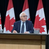 Le commissaire aux langues officielles dans une conférence de presse devant des drapeaux canadiens.