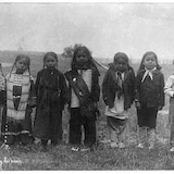 Seven small children stand in the grass. 