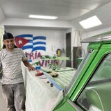 Un homme se tient à côté d'une voiture dans un garage. Derrière lui, un drapeau cubain est affiché sur le mur.